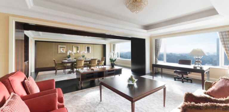 This image showcases an expansive and elegant living and dining space with a focus on luxury and comfort. The room is divided by a large opening framed by dark wood accents, leading from the cozy living area to a formal dining space. Both areas are adorned with plush chairs, a large dining table, and modern decor, exuding sophistication. The ceiling features intricate moldings, and the chandelier adds a touch of opulence. The large windows provide ample natural light, enhancing the space's open and airy feel. The overall design is a perfect blend of classic and modern styles.