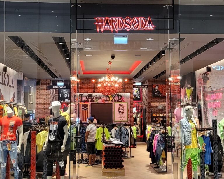 A vibrant clothing store named "HARDSODA" featuring mannequins dressed in bold, edgy fashion, with a neon-lit sign above the entrance and eclectic interior design elements, including red chandeliers, brick walls, and artistic posters.