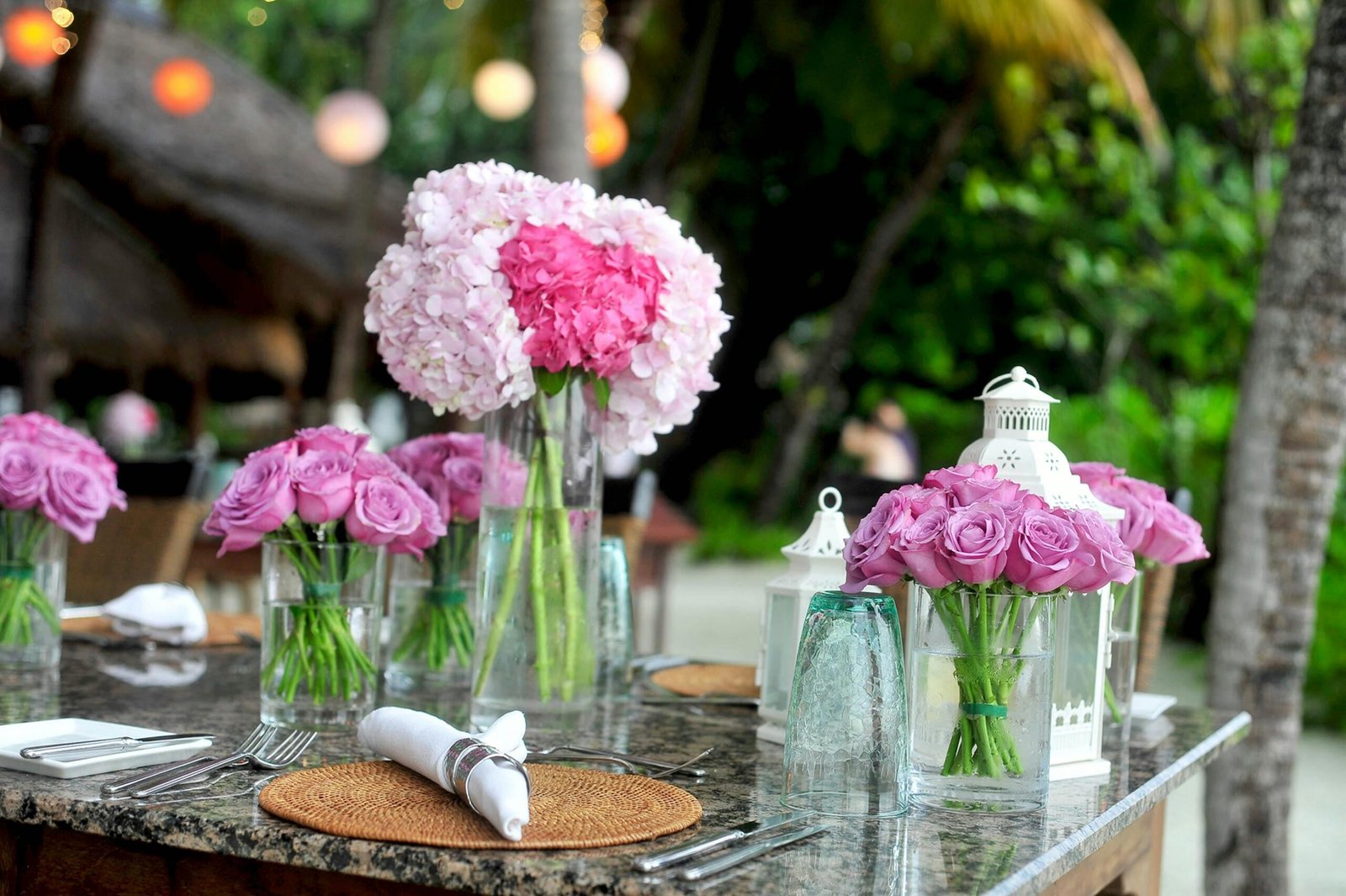 A beautifully decorated outdoor table with pink roses and hydrangeas in glass vases as centerpieces, set for a special event.